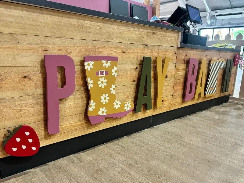 Colourful wooden sign spelling "PLAYBAR" mounted on wooden wall inside leisure venue, with red strawberry prop at base.