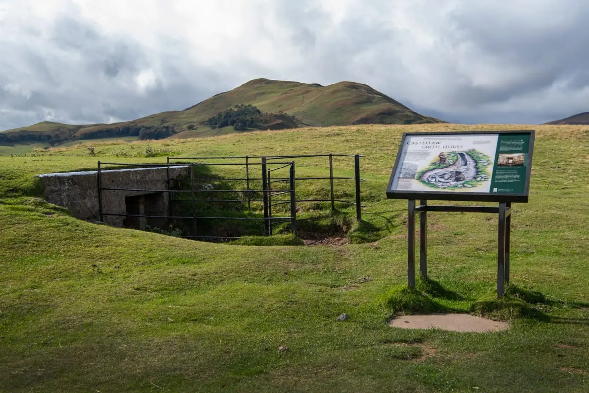 Archaeological site with stone ramparts, metal gates and an interpretive sign in rolling moorland beneath hills.