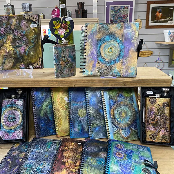 Display of decorative notebooks and journals with ornate patterned covers in jewel tones and metallics on wooden shelves in an art gallery shop.