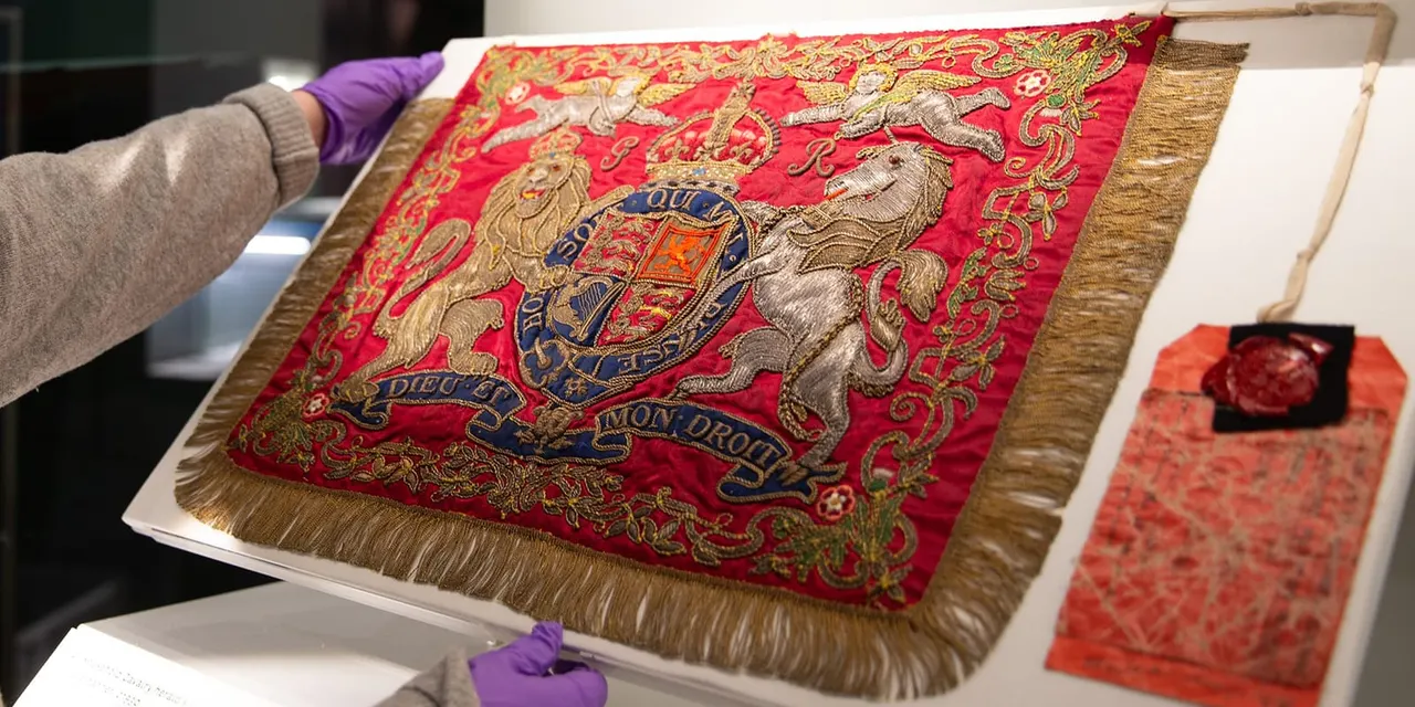 Person in purple gloves handling a red and gold embroidered royal coat of arms banner on display.