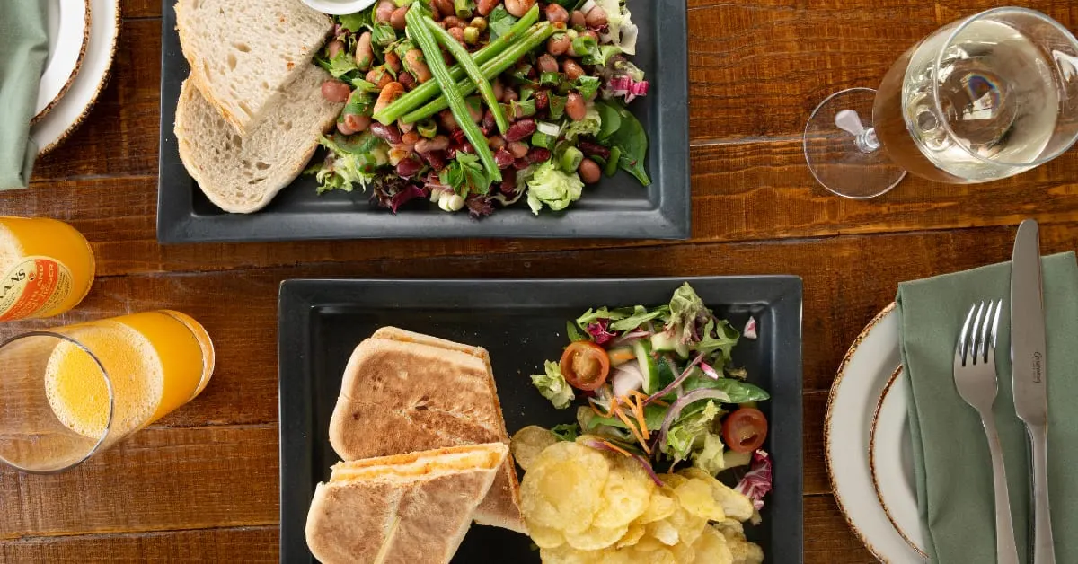 Overhead view of two plated meals with bread, salad, and crisps, alongside orange juice and water glasses on a wooden table.