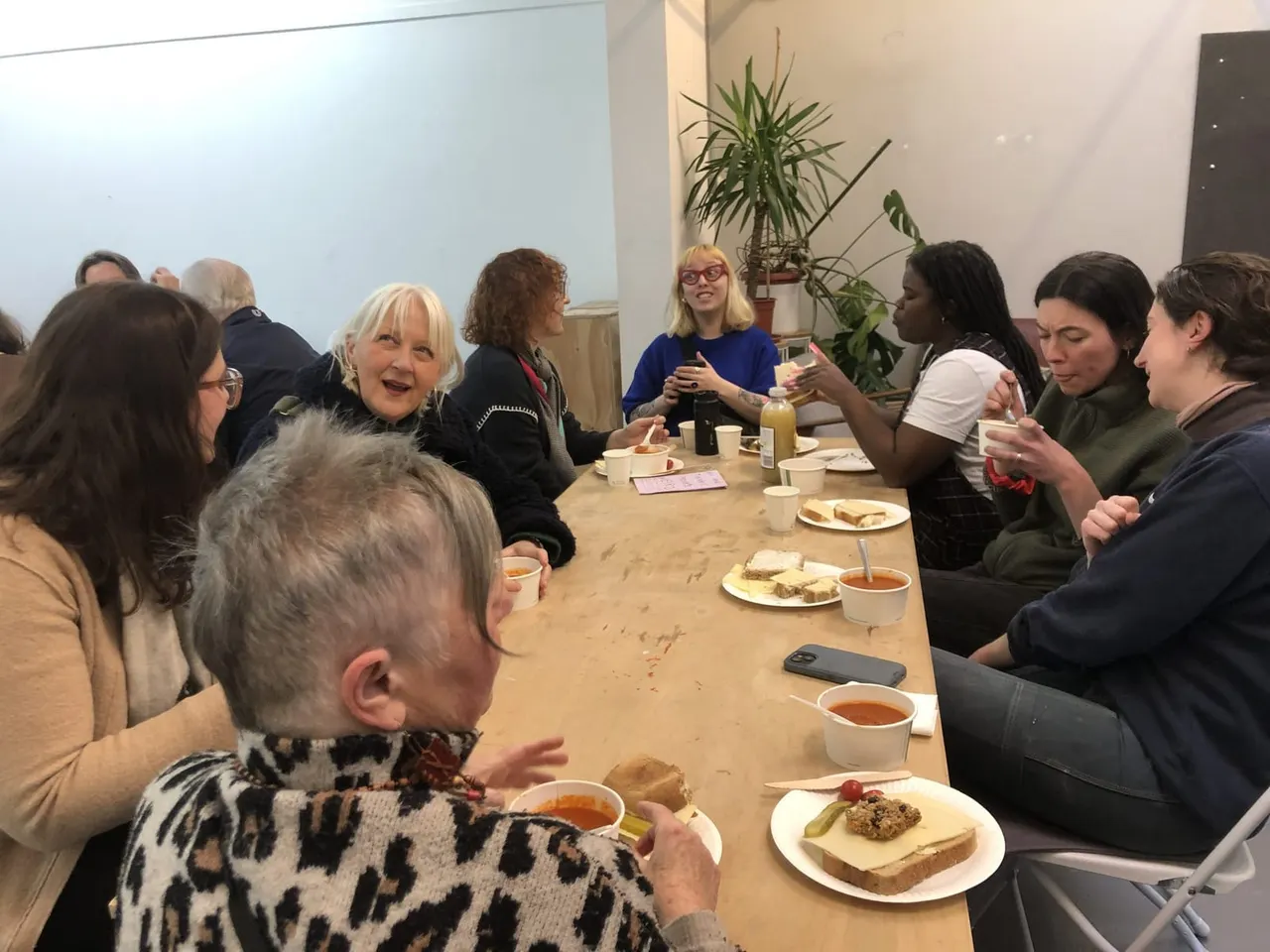 A group of people of mixed ages eating and chatting together around a wooden table with food and drinks in an indoor space with plants.