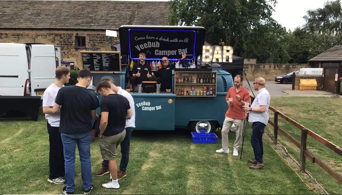 Visitors gather around a mobile camper bar serving drinks at an outdoor venue, with staff inside the converted van and guests standing on a grass area.
