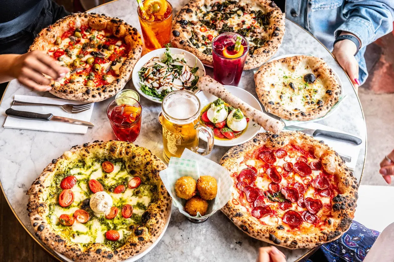 Multiple wood-fired pizzas with various toppings, drinks, and sides on a marble table shared by diners.