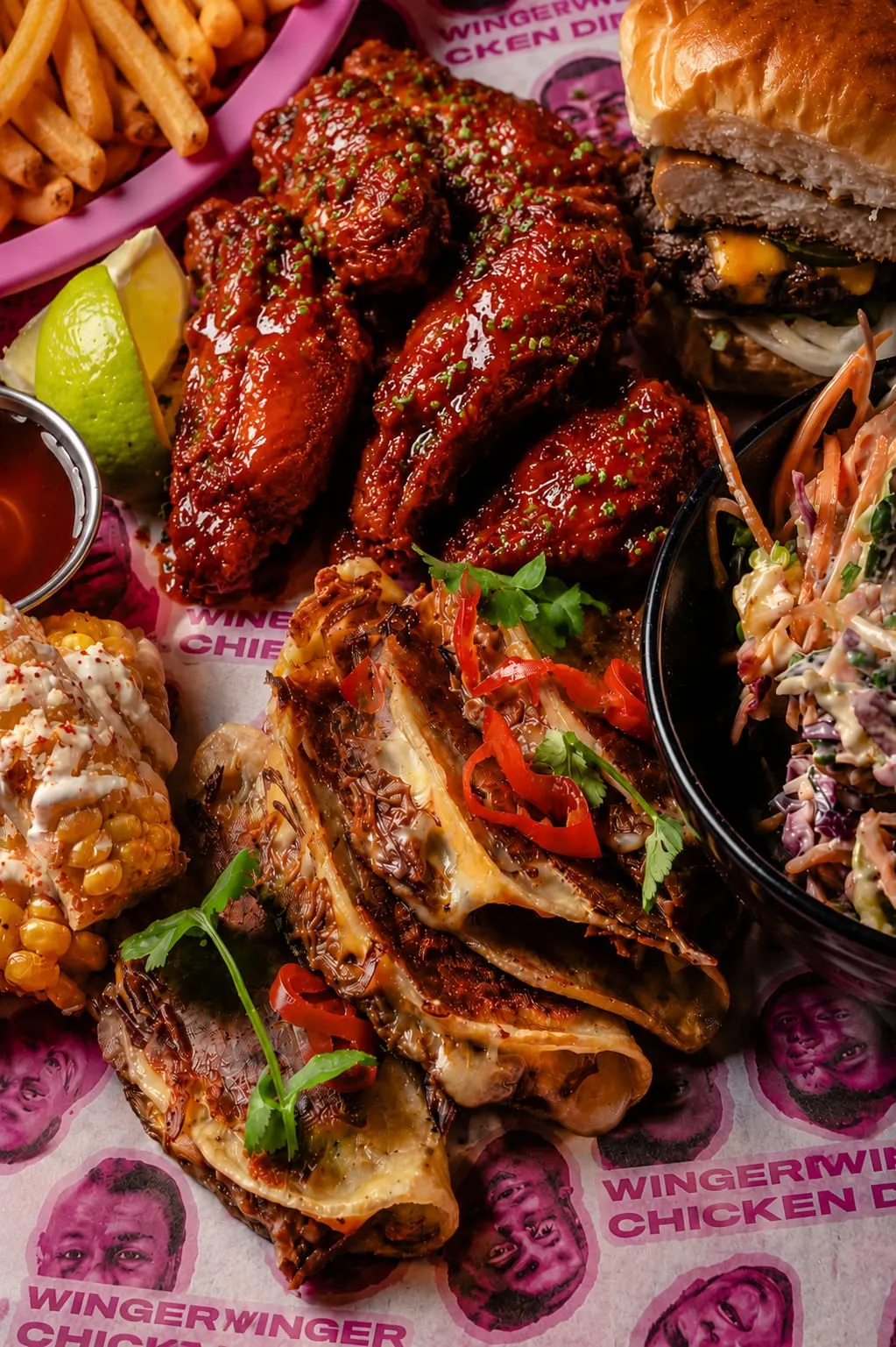 This is a PHOTOGRAPH.

Glazed chicken wings and drumsticks with fries, lime, burger, and coleslaw on branded paper.