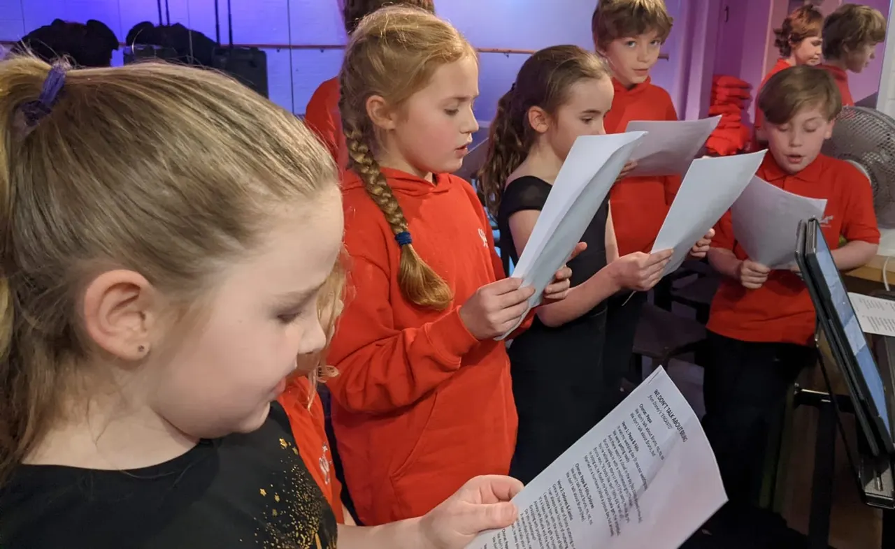 Children in red shirts holding scripts during a choir or performance rehearsal under purple stage lighting.