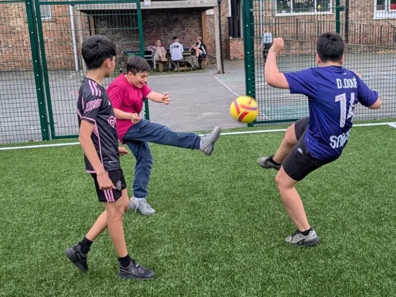 Young footballers playing on an indoor artificial pitch, one kicking a yellow ball whilst others defend.
