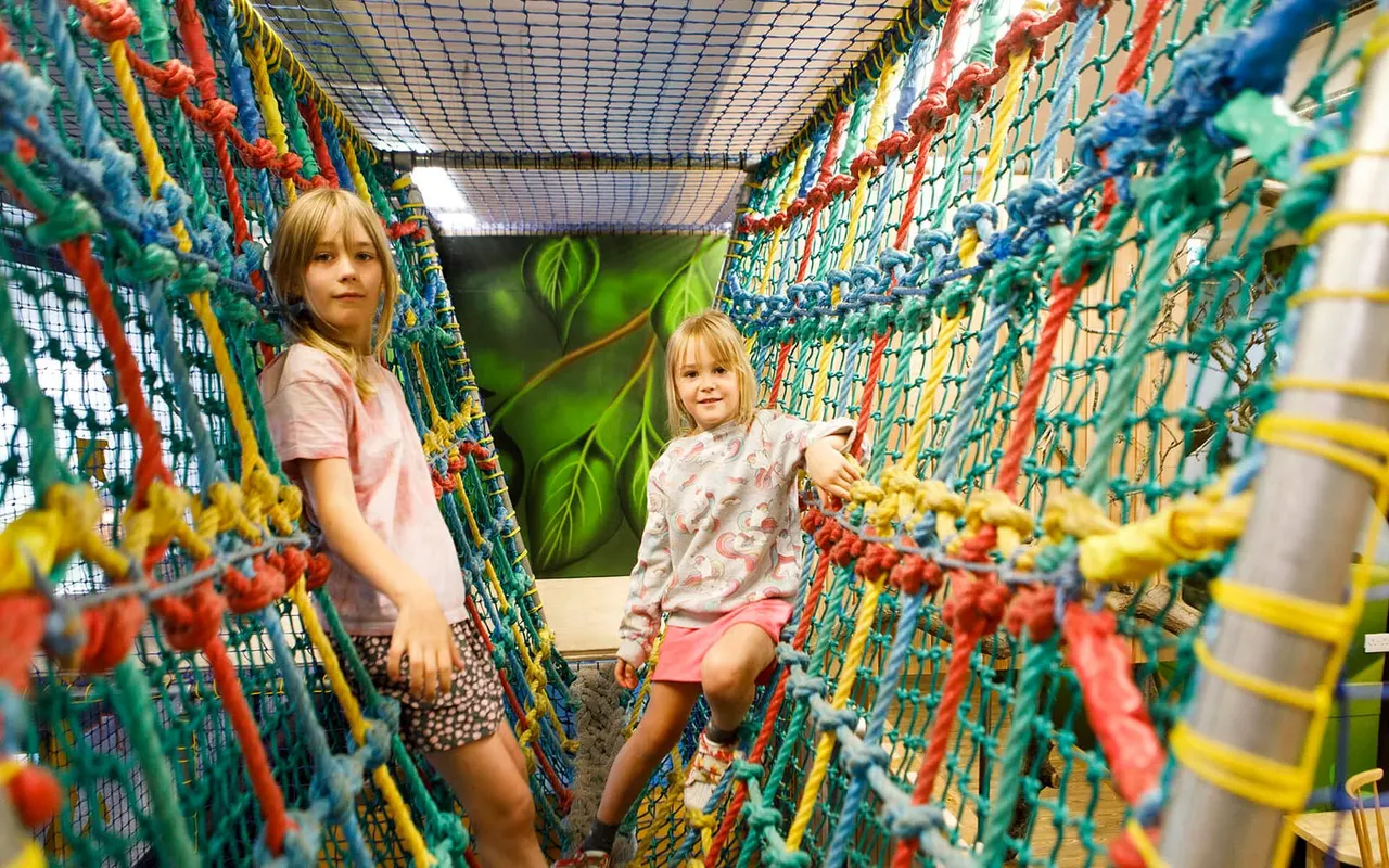 Two young children navigate through a colourful climbing net tunnel at an indoor play centre.