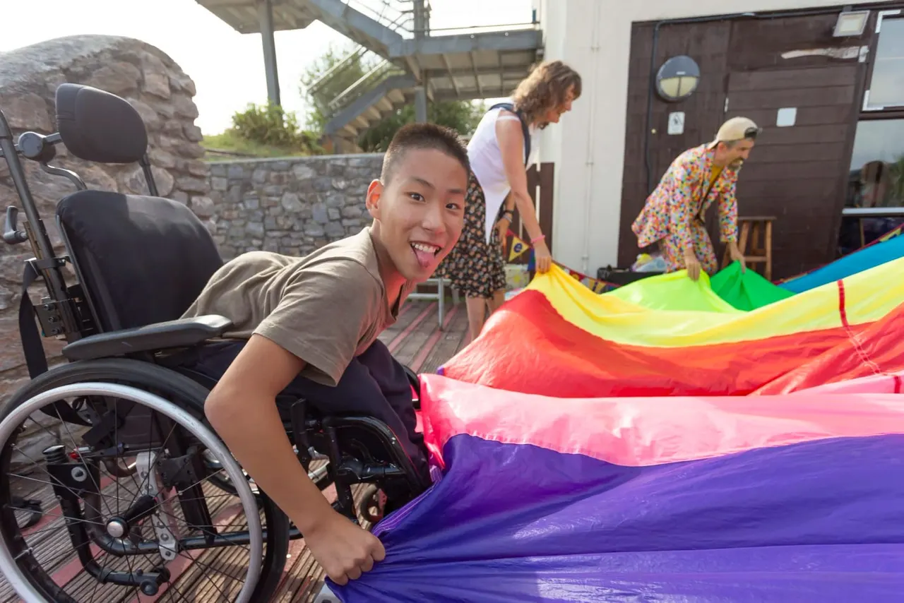 A young person in a wheelchair holds a colourful rainbow parachute during an outdoor group activity with other visitors and staff members.