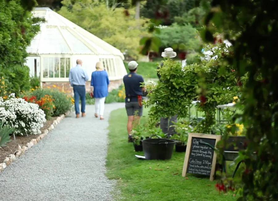 Three visitors walk along a garden path towards a white greenhouse, with flowering plants and a blackboard sign visible alongside.