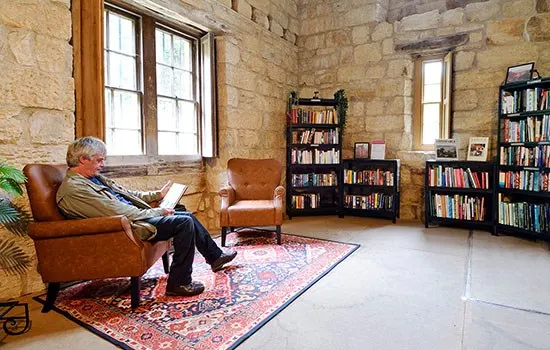 Older man sitting in leather armchair with laptop in stone-walled library with bookshelves and windows.