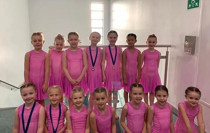 Group of young dancers in matching pink dresses posing in a dance studio, some wearing medals.