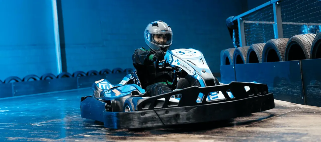 A person in a white and blue helmet drives a blue and white go-kart on an indoor track with protective barriers and tyres.