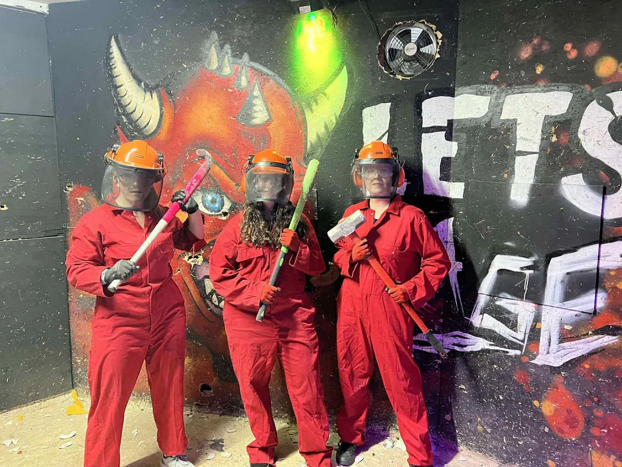 Four people in red jumpsuits and orange helmets holding foam weapons stand in front of a painted demon character backdrop.