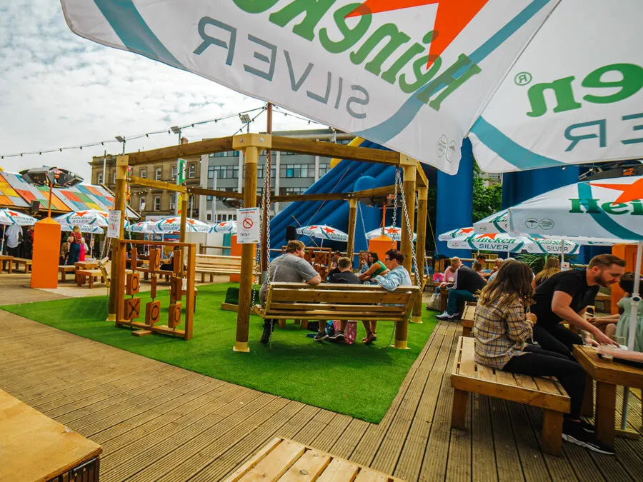 Outdoor festival seating area with visitors relaxing on wooden benches and swings under branded canopies on artificial grass.