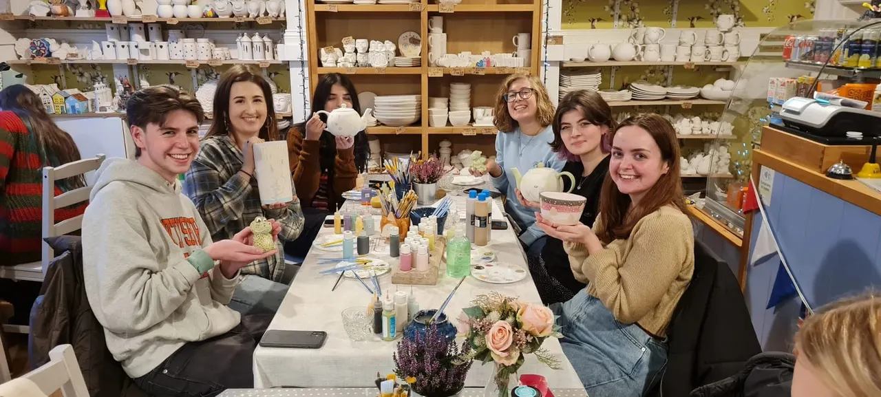 Group of young people decorating pottery at a workshop table surrounded by shelves of ceramics and painting supplies.
