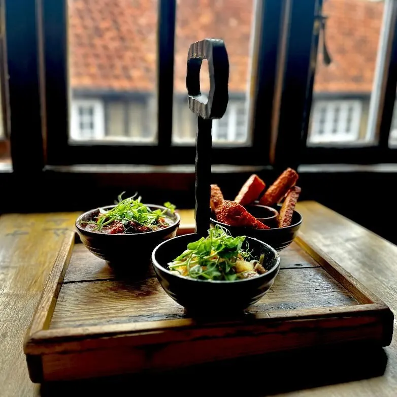 Tasting board with three small black bowls of food and crispy bread, displayed on wooden tray indoors with city views through windows behind.