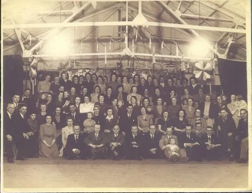 Large group photograph of formally dressed people gathered inside a wooden-beamed hall with overhead lighting.