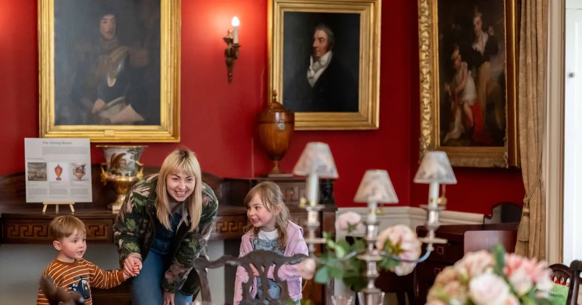 Woman and two young children exploring a grand red dining room decorated with period portraits, candelabras and ornaments.