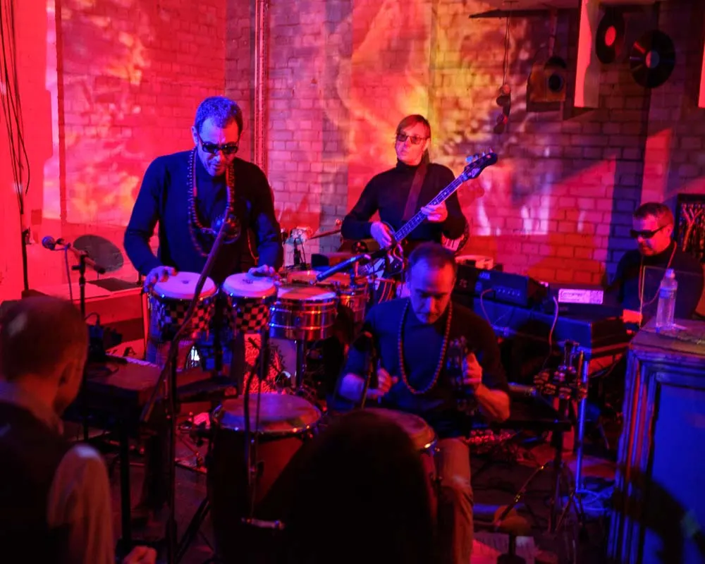 Band performing on stage with drums, bass guitar and keyboard under red and blue lighting, audience in foreground.