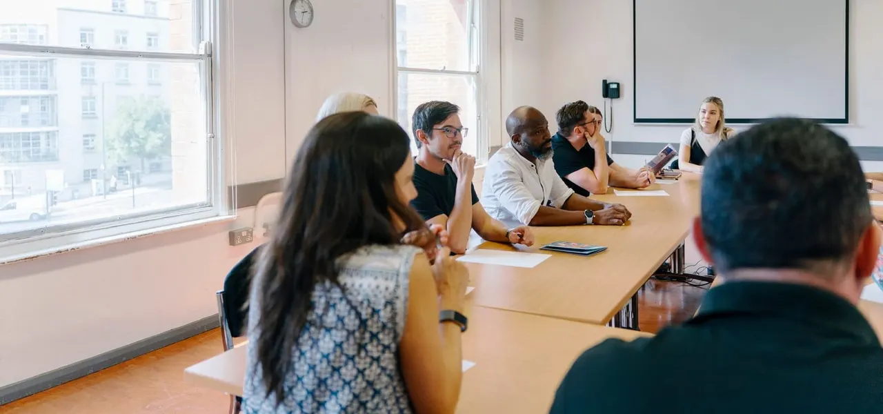 A group of people seated around a long table during a meeting or presentation in a bright, modern room with city views.