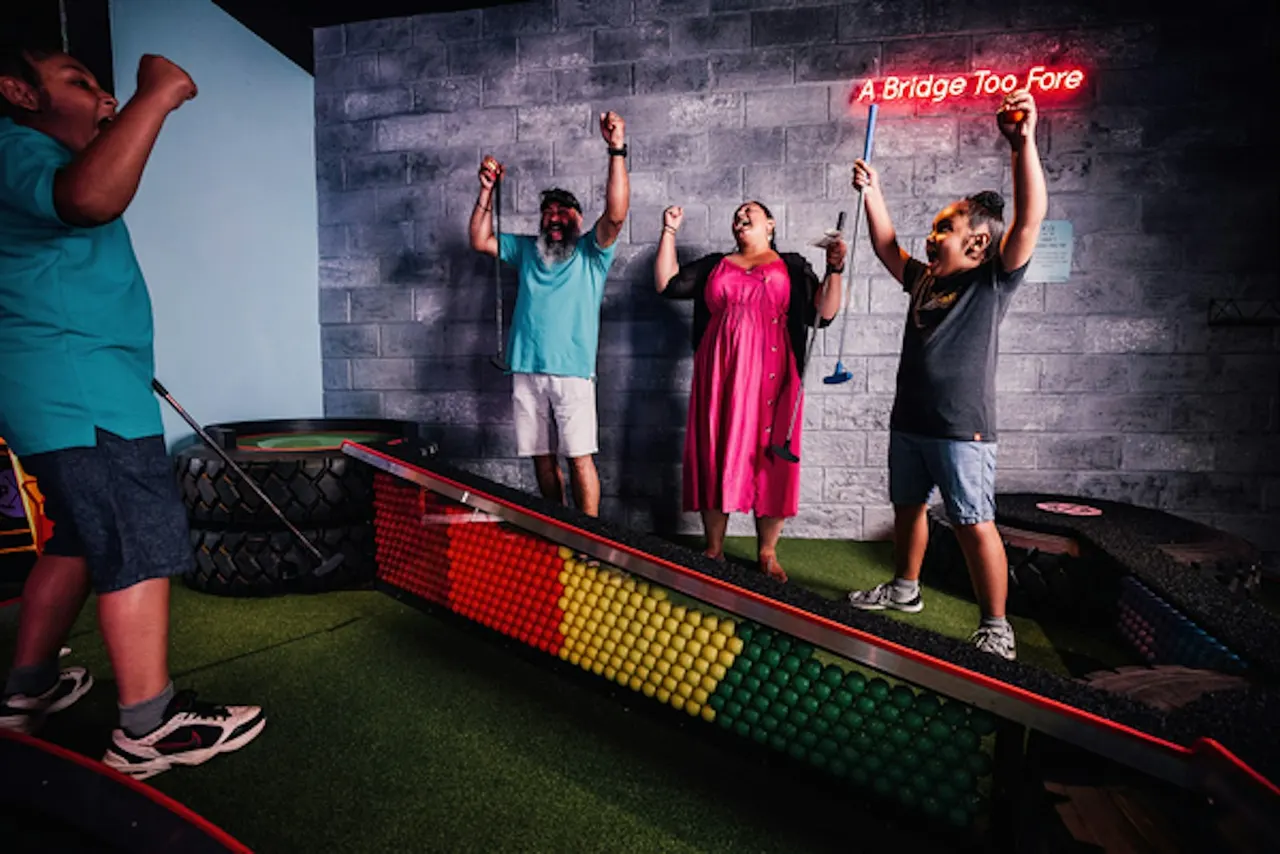 People playing crazy golf indoors, celebrating with arms raised near a coloured ball ramp and neon sign.