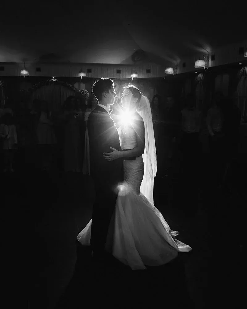 Bride and groom dancing together at night in a darkened venue, lit dramatically from behind.
