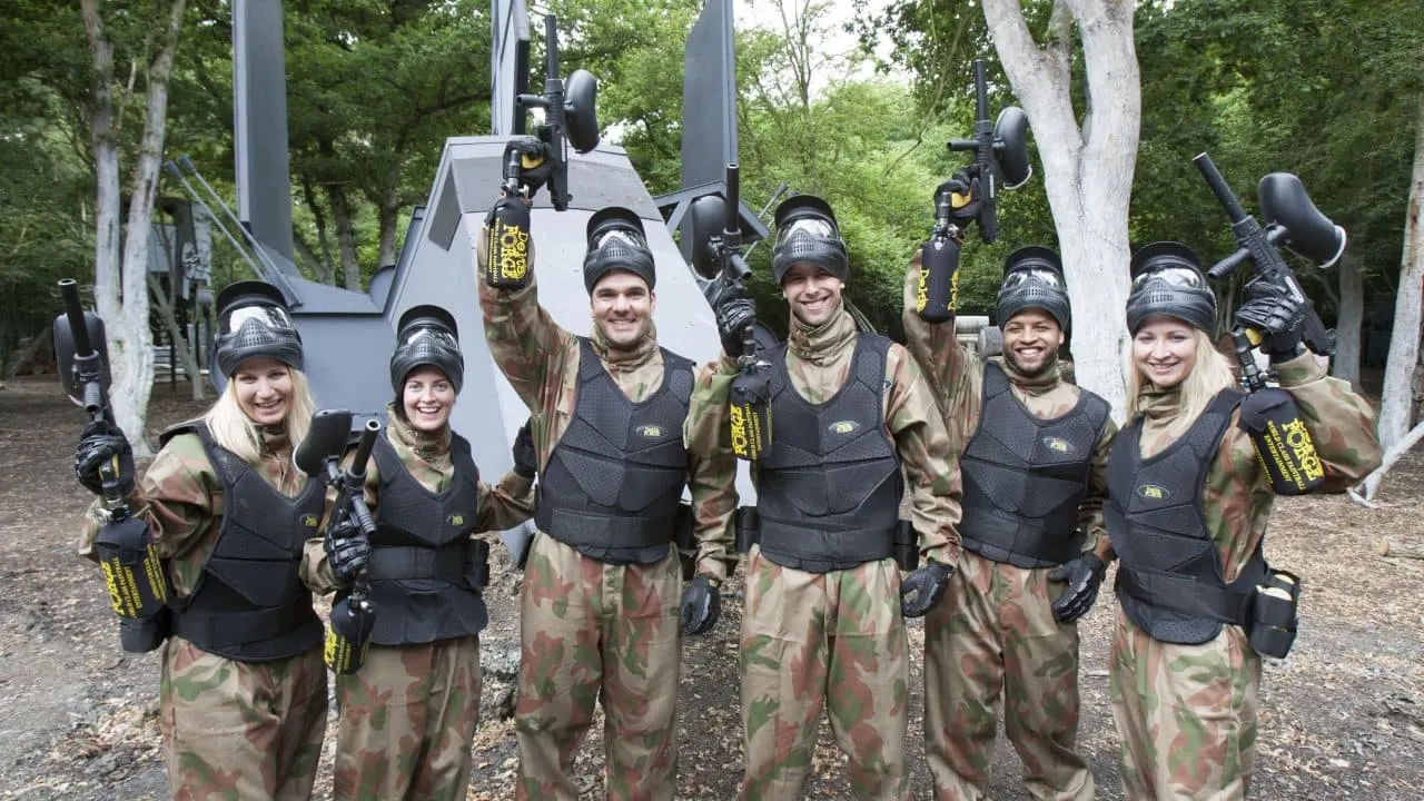 Group of seven paintball players in camouflage uniforms and protective gear posing together in a woodland arena.