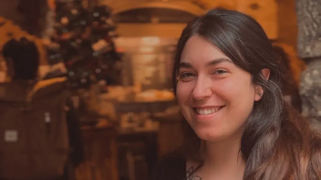 Young woman smiling at camera inside a warm-lit café with other patrons blurred in background.