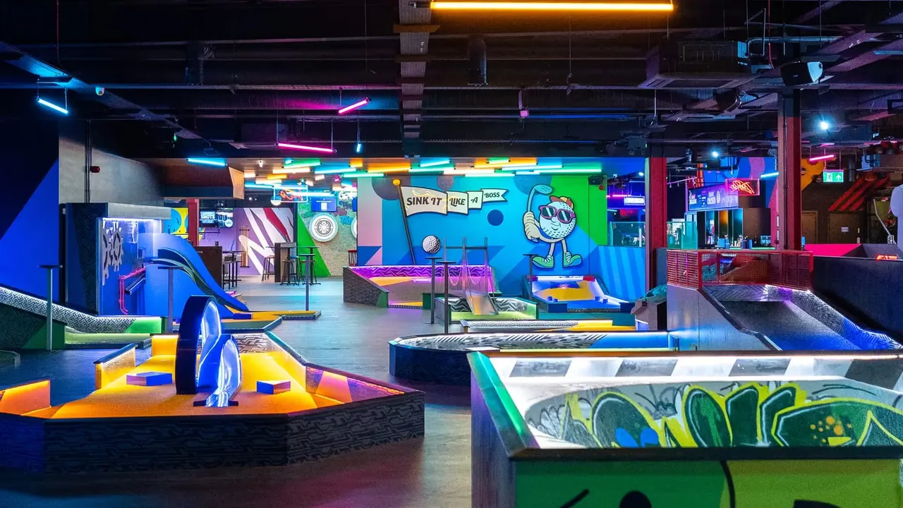 Brightly lit indoor mini-golf course with neon-coloured obstacles, cartoon wall art, and colourful neon tube lighting throughout the space.