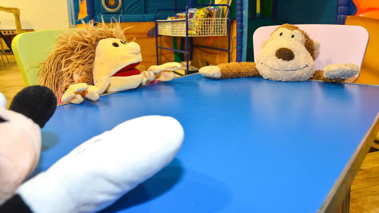 Soft toy puppets and characters interact at a blue table in a colourful children's play room.