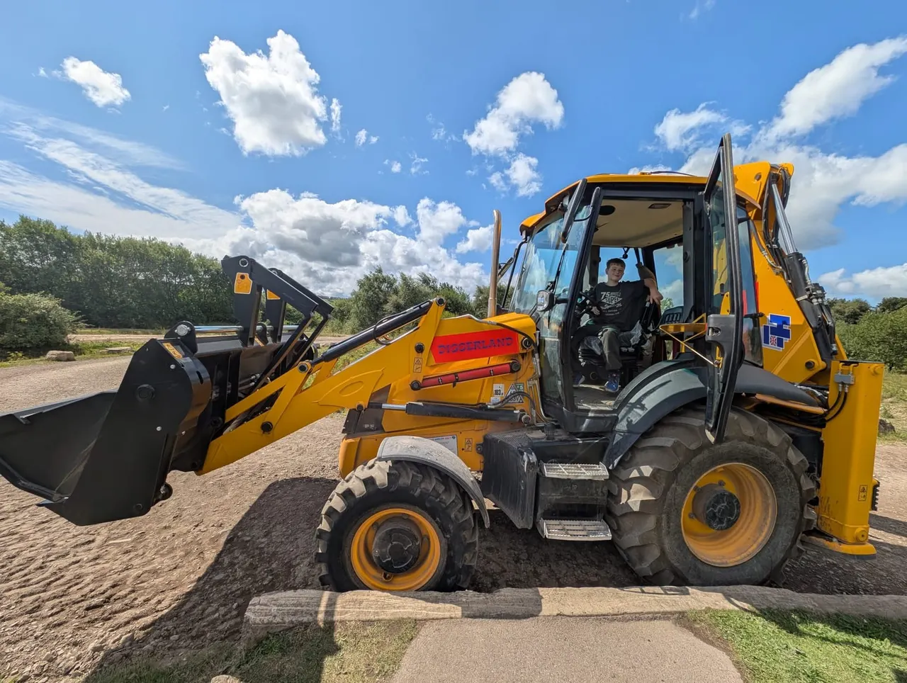 A person sits in the cabin of a yellow digger/telescopic handler with its bucket and boom arm raised against a blue sky.