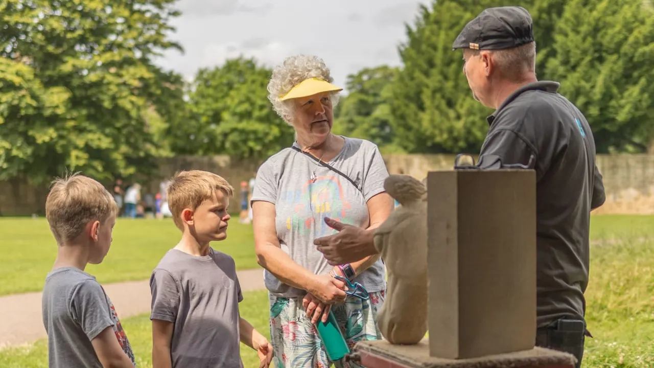 An adult guide talks to two young children and a small child beside stone ruins in a grassed area with trees and visitors in the background.