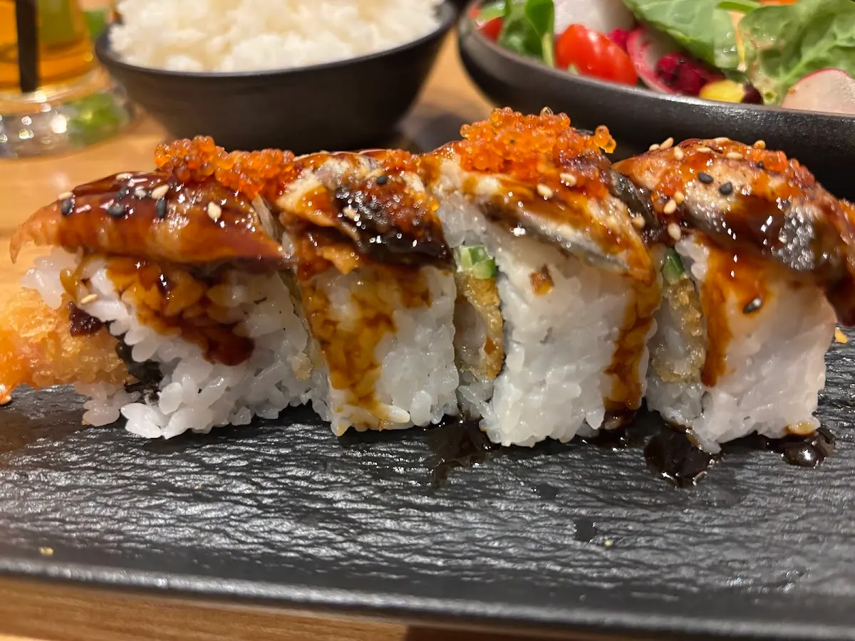 Glazed sushi rolls topped with seared fish and roe, served on a dark slate plate with rice and salad bowls behind.