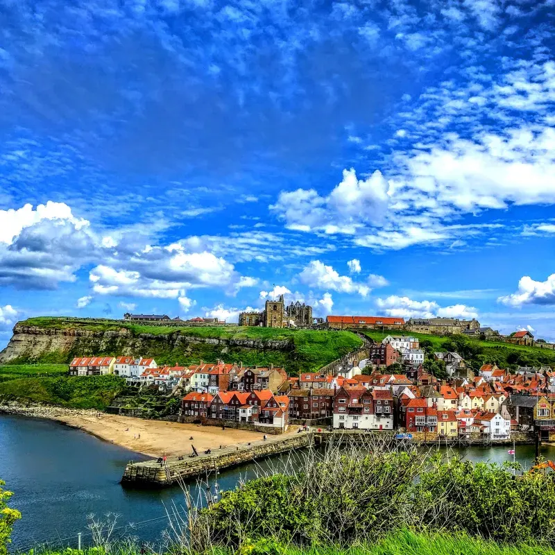 A seaside town with stone and terracotta-roofed buildings clustered around a harbour, backed by a green cliff topped with a ruined abbey.