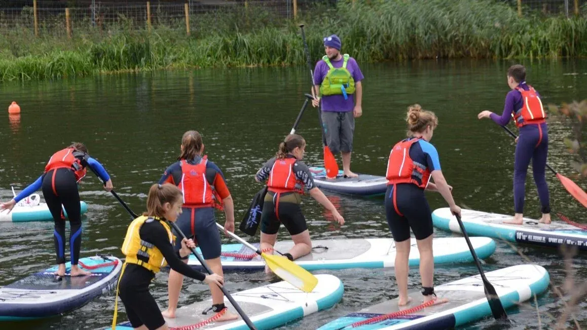 Children and instructors paddleboarding and learning water sports on a calm river with reeds along the banks.