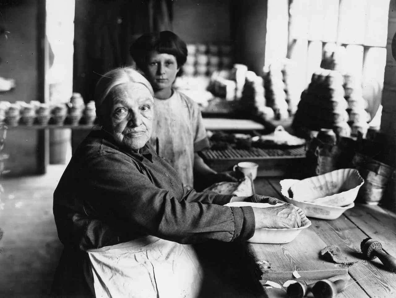 Elderly woman and younger woman working at a pottery table with clay vessels and tools in a workshop setting.