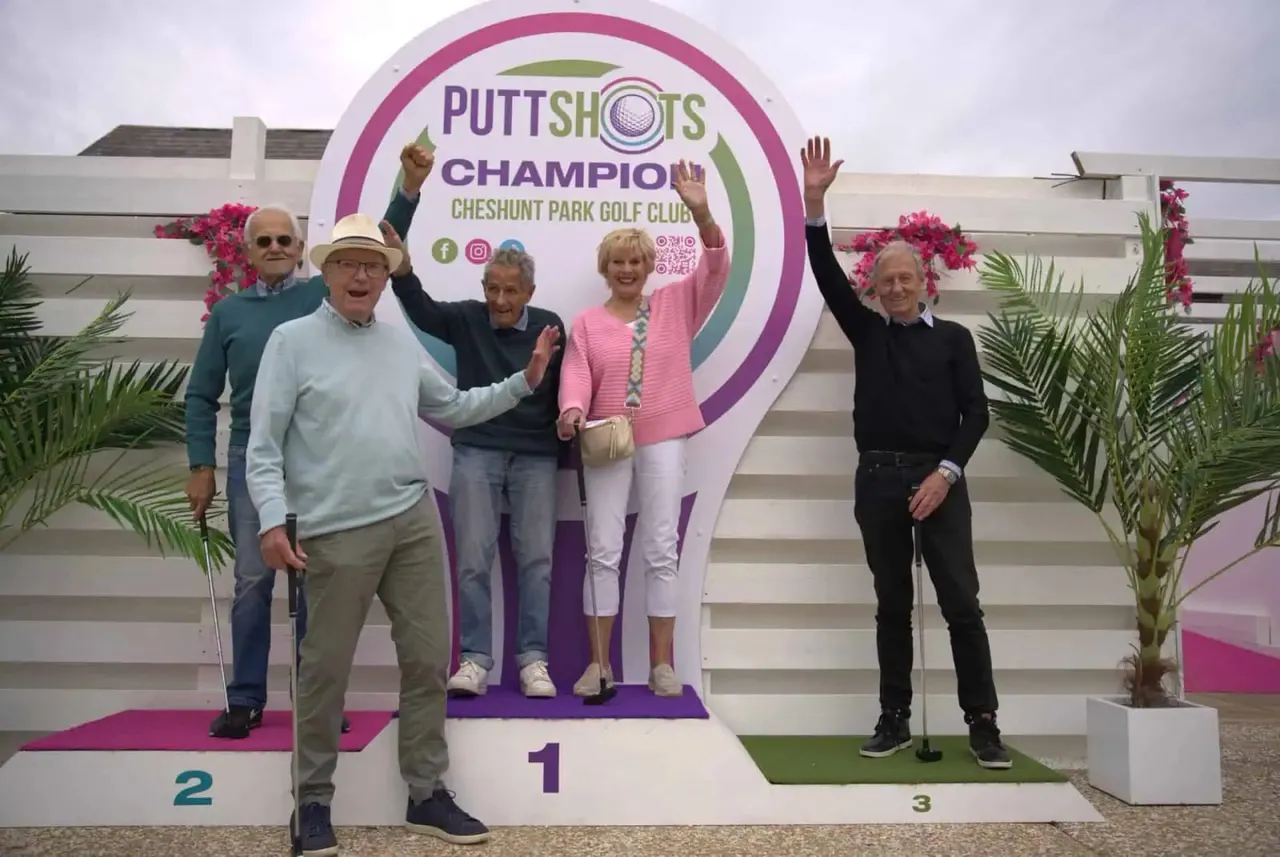 Five adults pose on a podium at a mini-golf venue, celebrating a putting championship with raised arms and smiles.