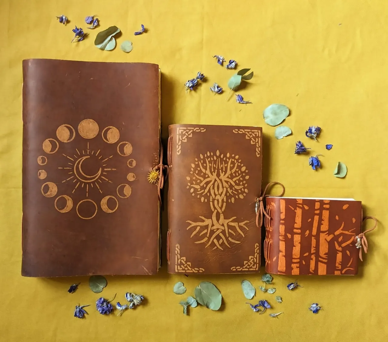 Three leather-bound journals with decorative embossing arranged on a yellow background, surrounded by dried flowers and leaves.