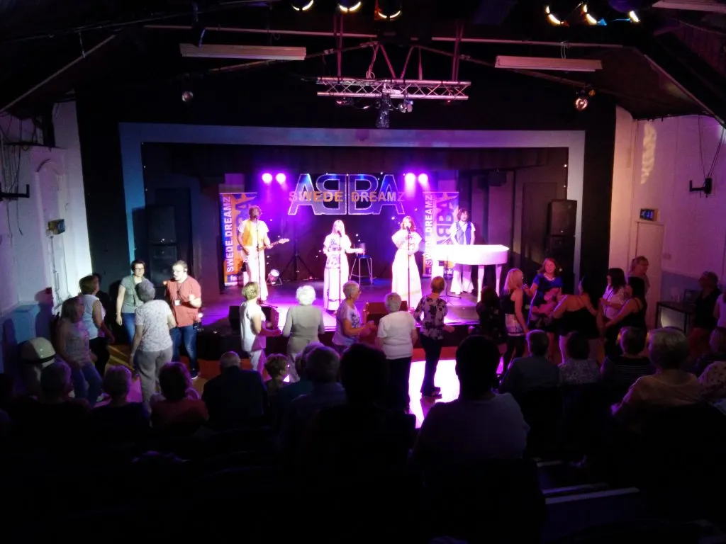 Live band performance on stage with purple lighting and audience members dancing and watching in an indoor venue.