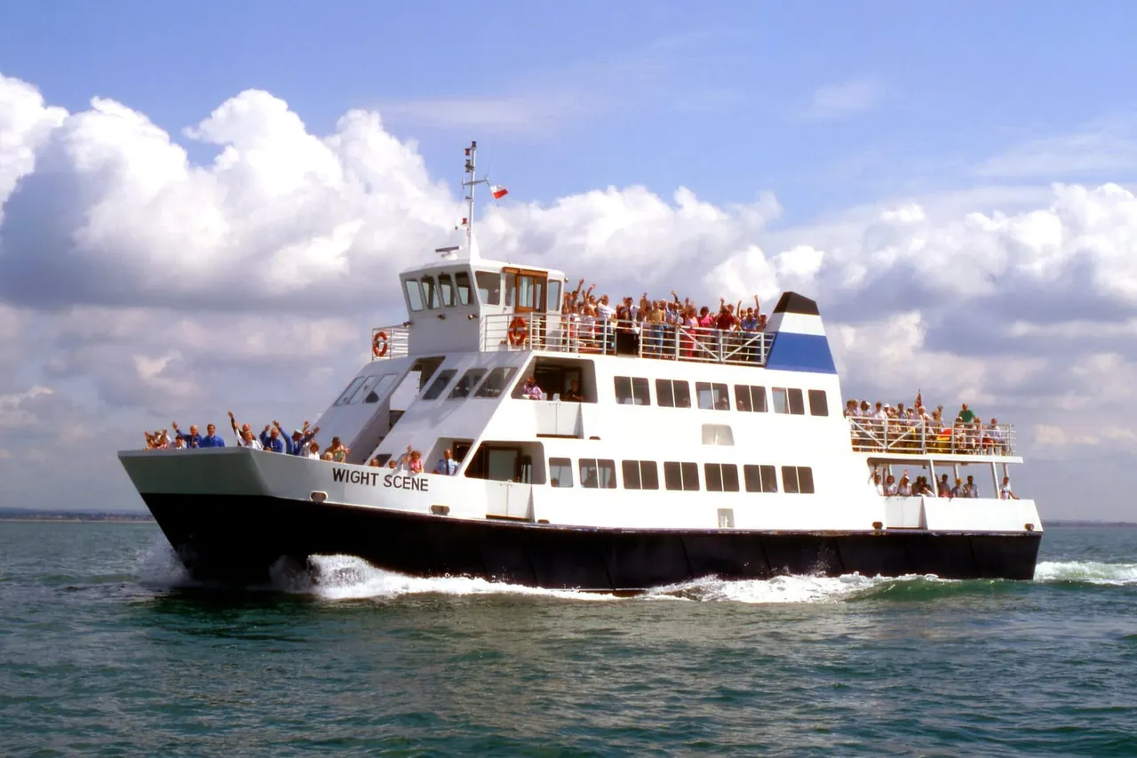 A white and black passenger ferry boat crowded with visitors on multiple decks, moving through calm sea waters under a blue sky with white clouds.