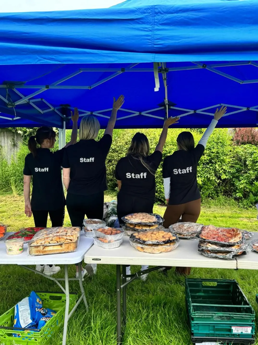 Staff members setting up a food service table under a blue marquee in a garden setting.