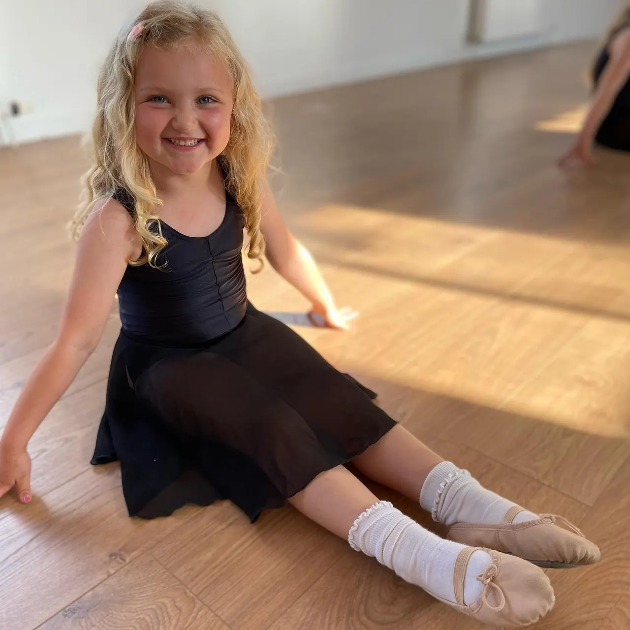 Young girl in black dance outfit and ballet pointe shoes sitting on wooden studio floor, smiling at camera.