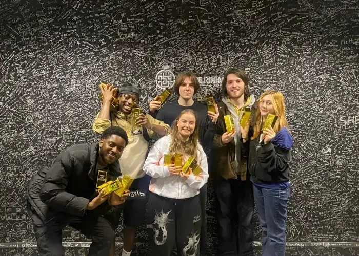 Group of six people holding gold trophies whilst posing together against a wall covered in handwritten text.
