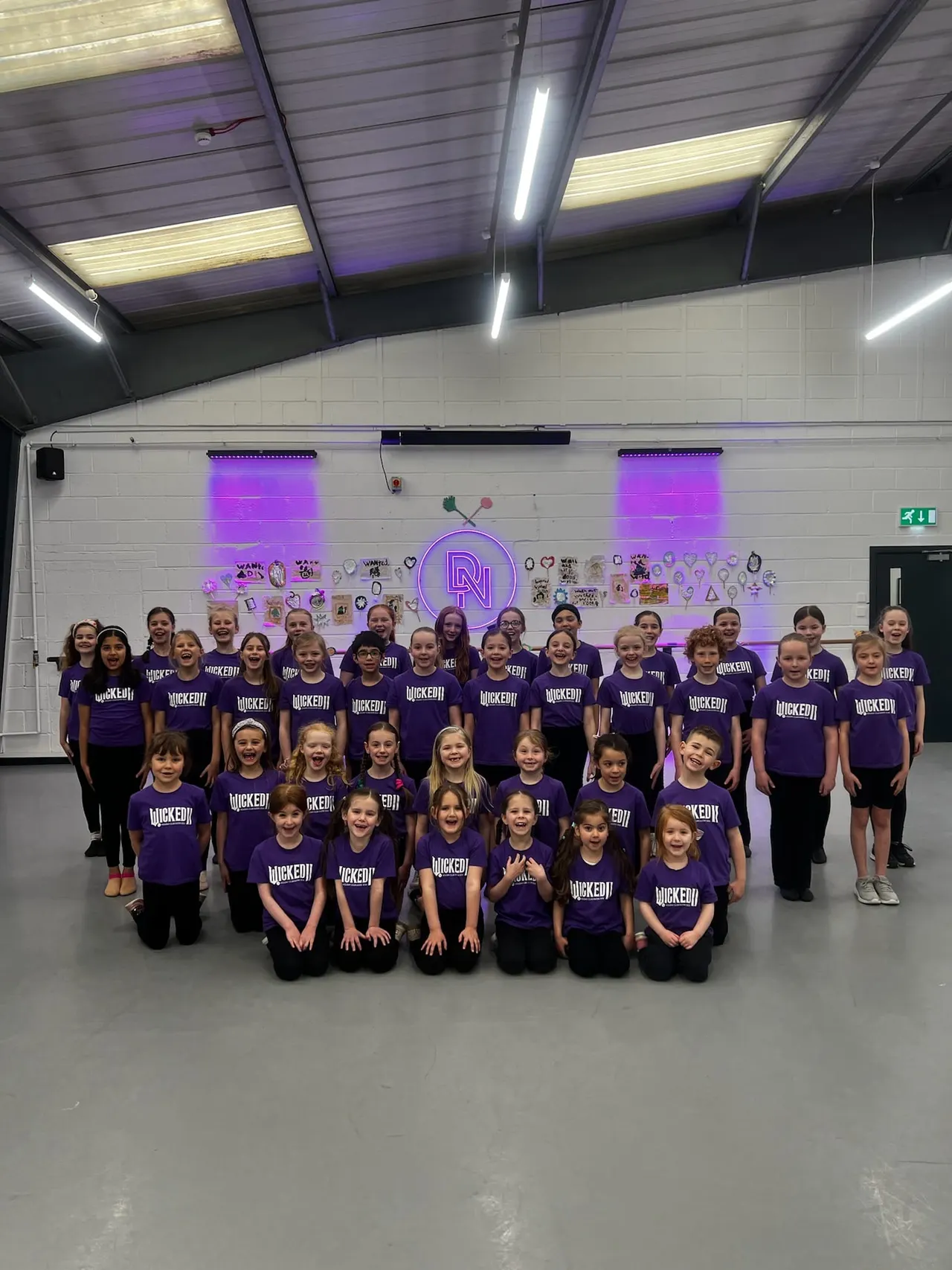 Large group of children wearing purple t-shirts posing together in a dance studio with purple neon lighting.