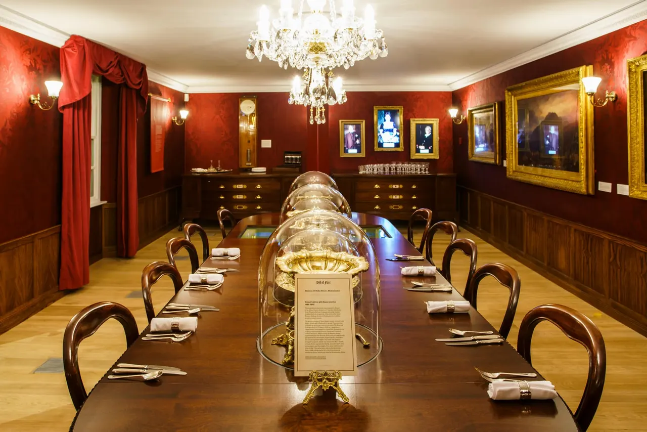 Elegant formal dining room with long table, place settings, crystal chandelier, red walls and gold-framed portraits.