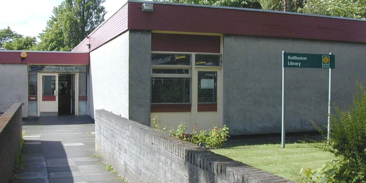 Modern single-storey library building with red roof and grey rendered walls, surrounded by trees and lawn.