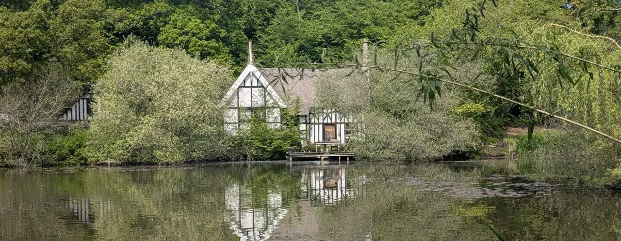 Tudor-style boathouse with black and white timber framing reflected in still water, surrounded by dense woodland and trees.