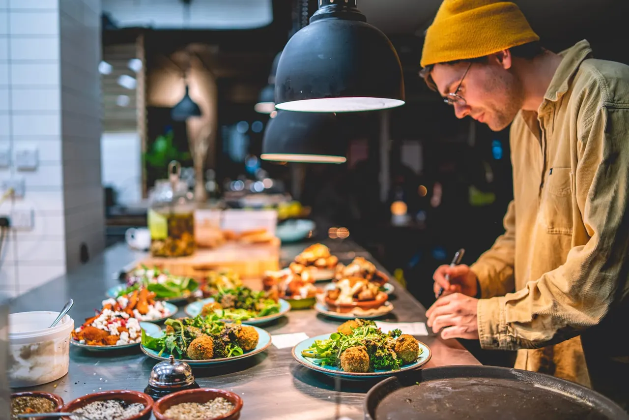 Chef plating up dishes with fried falafel and greens under a black pendant lamp in an open kitchen.