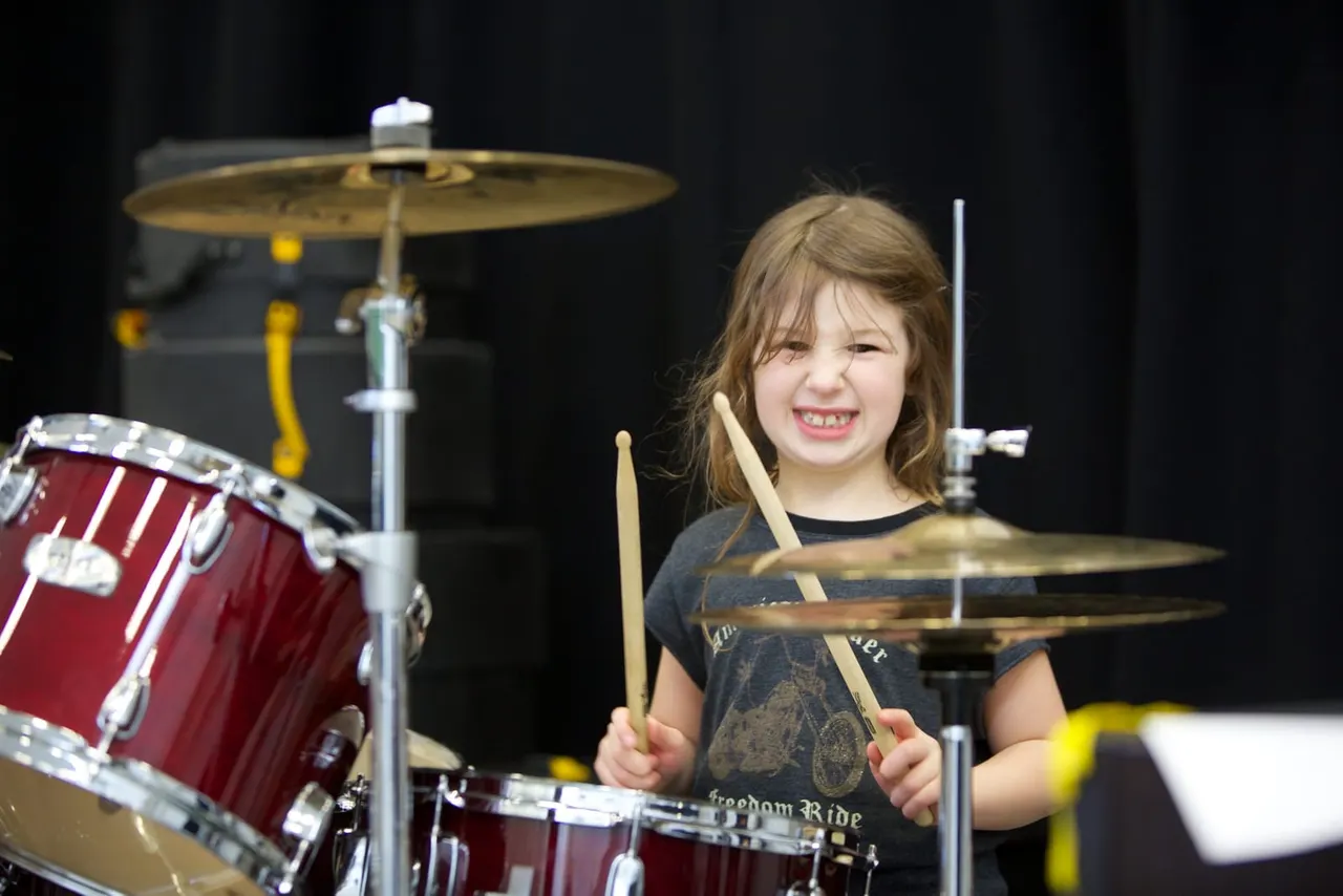 Young girl playing drums and smiling during a performance on stage with dark background.